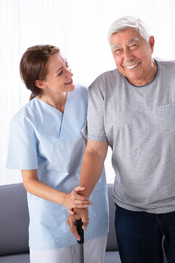 A female caregiver in scrubs assists an older man with gray hair, who is smiling and holding a cane, indoors.